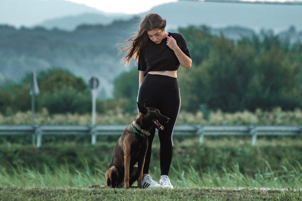 A woman in activewear stands outdoors with a German Shepherd on a grassy field.